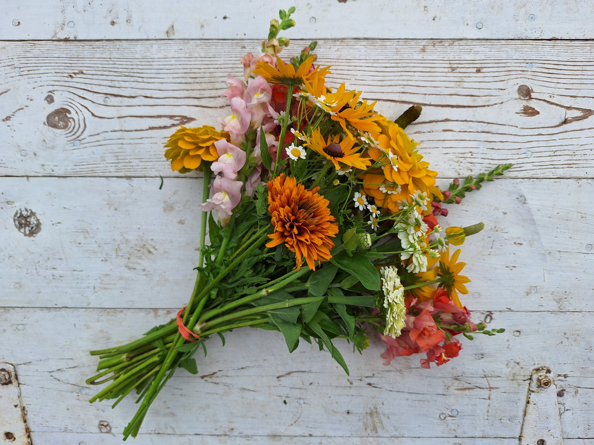 A bouquet of various flowers including snapdragons, cosmos, zinnia, and more, arranged together and wrapped in brown kraft paper, placed on a white wooden surface.